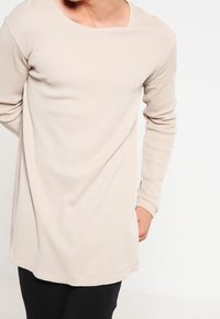 Beige långärmad tröja med en texturerad, ribbad design. Har en rundad halsringning och en något böjd nederkant, i kombination med svarta byxor.