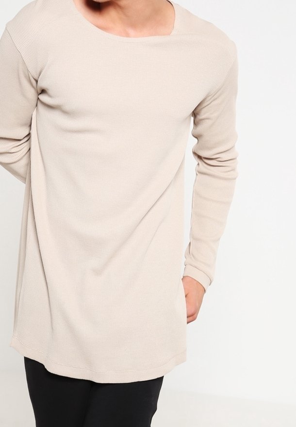 Maglia a maniche lunghe beige con un design a coste e texture. Presenta un colletto rotondo e un orlo leggermente curvato, abbinata a pantaloni neri.