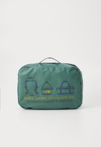 Green, quilted, rectangular backpack with carry handle. Features illustrations of backpack styles and descriptive text about compartments and materials.