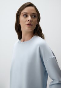 Jasnoniebieska bluza z okrągłym dekoltem, długimi rękawami oraz miękką, gładką fakturą. Posiada obniżone szwy na ramionach i minimalistyczny design.