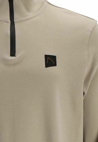 Beige Viertel-Reißverschluss-Pullover aus glattem Stoff, mit einem schwarzen Reißverschluss und einem kleinen schwarzen Patch mit einem orangefarbenen Design auf der Brust.