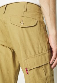 Béžové cargo kalhoty s texturovanou tkaninou, se zadní kapsou na knoflík a boční cargo kapsou, označené červeným logem.