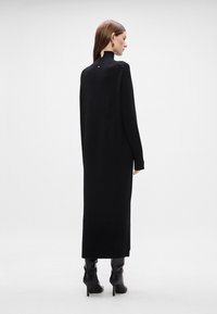 Μαύρο φόρεμα με μακριά μανίκια από μαλακό πλεκτό ύφασμα με ψηλό λαιμό, που διαθέτει πλαϊνές σχισμές και μινιμαλιστικό σχέδιο, συνδυασμένο με μαύρες μπότες με τακούνι.