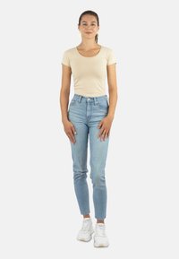 Beige eng anliegendes T-Shirt, hochgeschnittene hellblaue Jeans, Knöchellänge und weiße Sneakers. Das Modell steht mit den Händen an den Seiten vor einem schlichten Hintergrund.