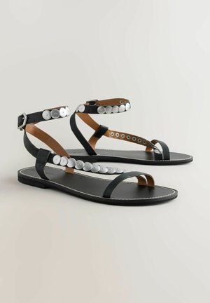 Sandales plates noires avec une finition en cuir lisse, dotées d'accents en bouton argenté le long des sangles et d'un design minimaliste.