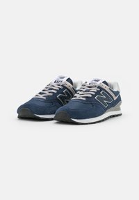 Navy sininen New Balance -lenkkarit, joissa on mokkanahka- ja verkko-yläosa, heijastavat yksityiskohdat, beige nauhat ja musta kumipohja. Malli 574.