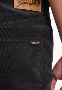 Un pantalon en velours côtelé noir présente une étiquette en cuir avec "Volcom" et une texture texturée, contrastée par un petit logo sur la poche.