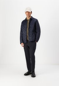 Jachetă din material de marină, quilted, cu nasturi, polo oliv dedesubt, pantaloni cargo negri și pantofi negri. Poartă o pălărie crem. Fundal simplu.