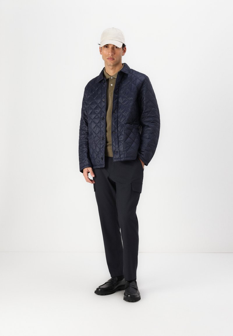 Veste matelassée bleu marine avec des boutons, polo olive en dessous, pantalon cargo noir et chaussures noires. Portant une casquette crème. Fond simple.