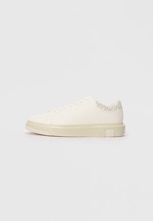 Sneaker bianco con tomaia in tela liscia, lacci piatti, colletto imbottito e un accento grigio testurizzato sul retro. Suola in gomma spessa.