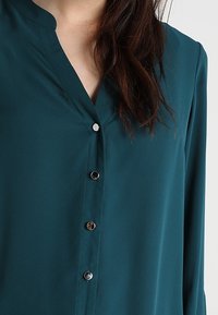 Turkos button-up blus med v-ringad krage, mjukt tyg och metallknappar. Har en avslappnad passform och långa ärmar.