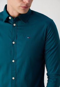 Chemise boutonnée couleur sarcelle avec col classique, boutons blancs et petit logo brodé en rouge, blanc et bleu sur la poitrine.