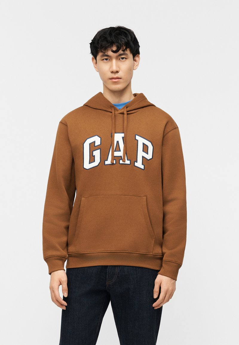 Νεαρός άνδρας με μαύρα σγουρά μαλλιά, φοράει καφέ φούτερ GAP και σκούρο τζιν, στέκεται μπροστά σε έναν απλό φωτεινό φόντο.