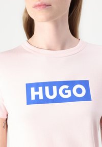 Lyserød bomulds t-shirt med et blåt rektangulært logo med hvid text "HUGO." Rund halsudskæring og korte ærmer.