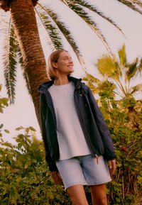 Jonge vrouw in spijkershorts en een donker jack die lacht, staand tussen groene planten en palmbomen bij zonsondergang.