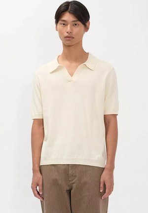Joven con el cabello oscuro y liso, vestido con una camisa polo crema de manga corta y pantalones marrones sobre un fondo claro y liso.