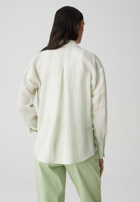 Camicia a maniche lunghe verde chiaro, con texture e piega sul retro, indossata su pantaloni larghi verde chiaro. Vestibilità morbida e rilassata.