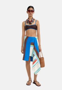 Brązowy stanik bikini z niebieskimi wykończeniami, kolorowa spódnica w kwiaty z niebieskimi, zielonymi i brązowymi paskami, brązowe sandały i pleciona torba.