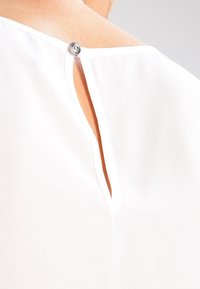 Blusa bianca con scollo rotondo e chiusura a bottone. Presenta un piccolo apertura a chiave sul retro, che mette in risalto la texture liscia.