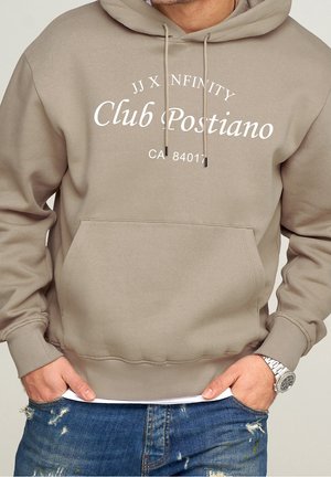 Sweat à capuche beige avec le texte « JJ X NFINITY Club Postiano CA 84011 », porté par une personne avec les mains dans les poches d’un jean bleu usé et une montre argentée.