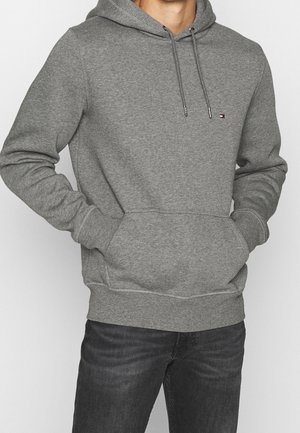 Mann trägt ein graues Kapuzen-Sweatshirt mit Fronttasche und kleinem Logo auf der linken Brust, kombiniert mit dunklen Jeans.