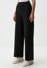 Pantaloni a gamba larga in tessuto a coste grigio scuro con vita elasticizzata e tasche laterali, abbinati a scarpe da ginnastica bianche su uno sfondo neutro.