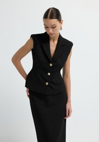 Blazer noir sans manches sur mesure avec des boutons dorés, épaules structurées et deux poches avant, associé à une jupe noire ajustée.