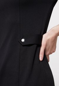 Musta mekko, jossa on sivutasku, suljettuna nepparilla. Kangas on sileä ja mattapintainen. Lähikuva kädestä, joka lepää taskussa.