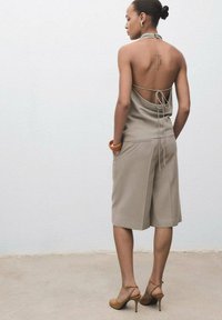 Beige halterneck-top med åben ryg, løse shorts og højhælede sko. Bemærkelsesværdige detaljer inkluderer en sløjfe i ryggen og en ryg-tatovering.