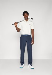 Αθλητική ενδυμασία γκολφ με ανοιχτό μπεζ πόλο μπλουζάκι με λογότυπο, ναυτικές παντελόνες και λευκά παπούτσια γκολφ με μπλε λεπτομέρειες. Ο γκολφέρ κρατά ένα ραβδί.