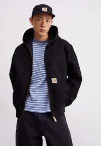 Melna zip-up kapuce jaku ar auklu, kurai ir priekšējais kabata un logotipa uzlīme. Zem tās ir svītraina zila un baltā krekls. Tumši bikses.