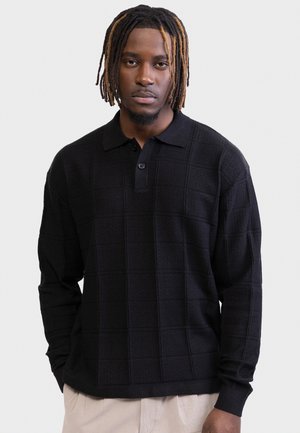 Άνδρας με dreadlocks μέχρι τους ώμους φοράει μαύρο, υφασμάτινο μακρυμάνικο πόλο και ανοιχτόχρωμο παντελόνι, στέκεται μπροστά σε απλό φόντο.