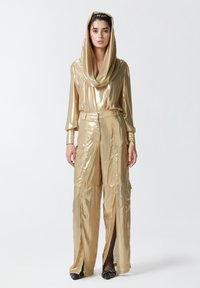 Metalni zlatni outfit s dugim rukavima, kapuljačom i širokim hlačama s prorezima. Tkanina ima glatku, sjajnu teksturu.