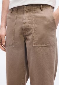 Μπεζ βαμβακερό παντελόνι με μεγάλες ορθογώνιες μπροστινές τσέπες, κλείσιμο με κουμπί και ένα χέρι μερικώς στην δεξιά τσέπη.