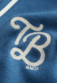 Modrá textília obsahuje výrazné biele vyšívané logo so stylizovanými písmenami "TB" a "BAKER." Tkanina vykazuje textúru a hĺbku.