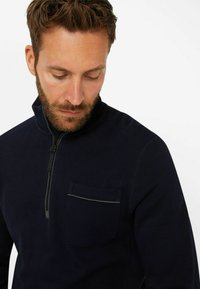 Marinefarbener Reißverschluss-Pullover aus strukturiertem Stoff, ausgestattet mit einer Brusttasche mit kontrastierendem Streifen. Eng anliegender Kragen und schlankes Design.
