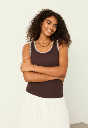 Femeie cu păr creț, purtând un top maro fără mâneci cu tiv alb și o fustă albă, zâmbind cu brațele încrucișate.