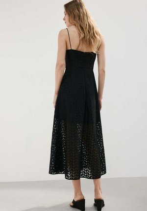 Kobieta o długich falowanych włosach stoi tyłem, ubrana w bezrękawnikową czarną koronkową sukienkę midi i czarne sandały na koturnie.