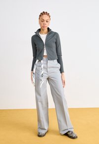 Veste gris zippée, design crop, assortie à un pantalon large gris. Écharpe blanche à motif noir nouée à la taille, chaussures à rayures. Fond jaune.