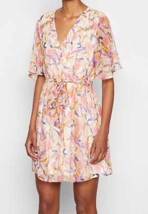 Kobieta ubrana w krótką sukienkę w kwiaty z falbanowymi rękawami i paskiem wiązanym w talii w pastelowych odcieniach różu, żółci i fioletu.