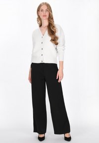 Fehér kötött kardigán V-nyakkal, gombos előlappal és bordázott mandzsettával, fekete bő szárú nadrággal és fekete magassarkú cipővel párosítva.