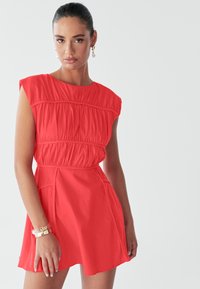 Modelka oblečená v jasne červenom šate s krátkymi rukávmi a zberanými detailmi, skombinovaná so zlatými náramkami a elegantným účesom na jednoliatom pozadí.