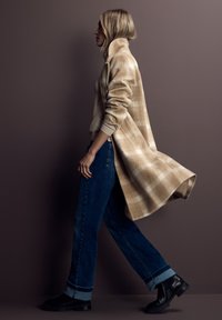 Cappotto lungo a quadri beige con tessuto texturizzato, indossato sopra un maglione chiaro, abbinato a jeans blu scuro con risvolto e stivaletti neri.