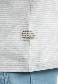 Tela de rayas grises con una etiqueta en el dobladillo que dice "Petrol Ind". La etiqueta es de color crema con texto azul y negro, cosida al tejido.