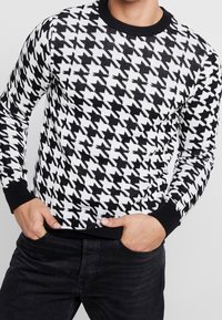 Černobílý pletený svetr se vzorem houndstooth, žebrovanou strukturou a černým kulatým výstřihem a manžetami. Nosený s černými kalhotami.