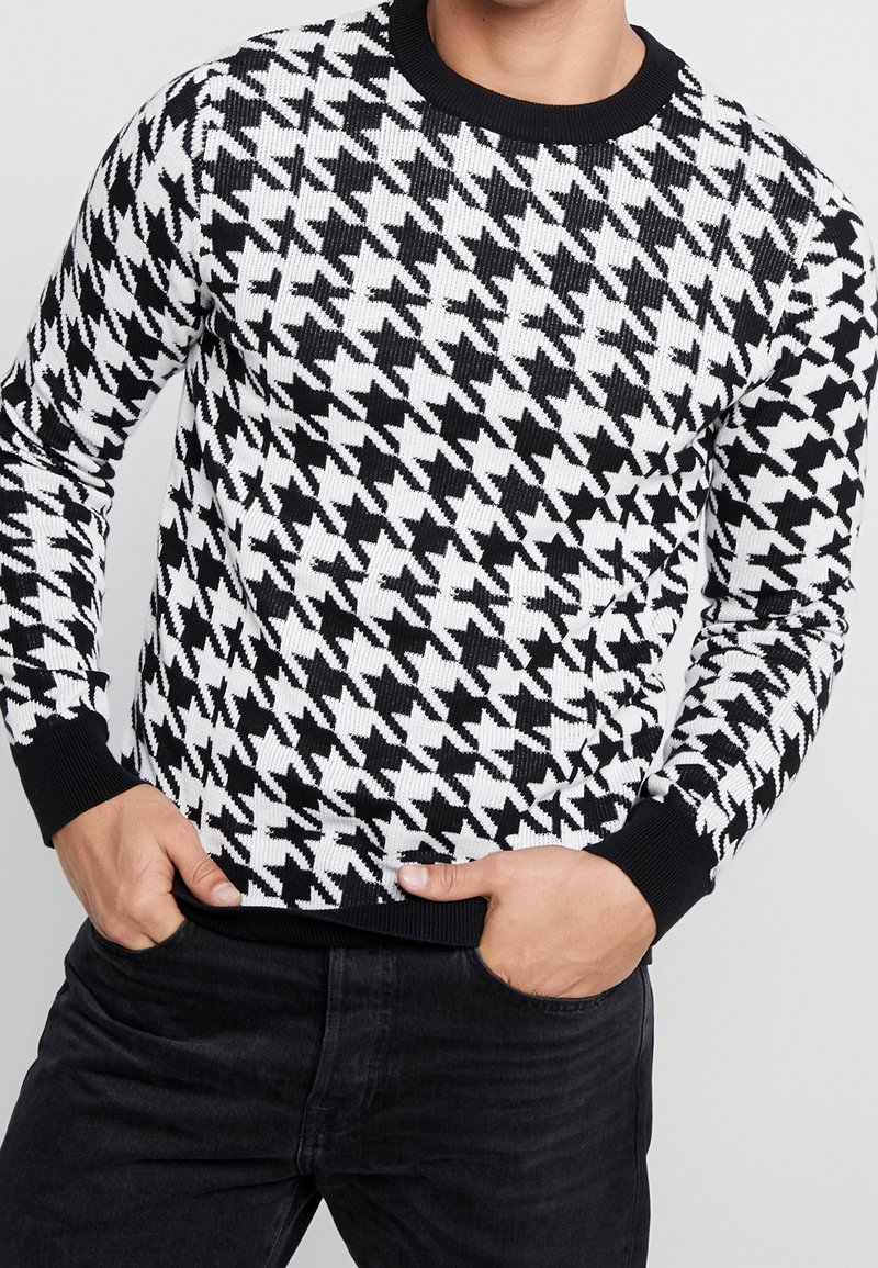 Černobílý pletený svetr se vzorem houndstooth, žebrovanou strukturou a černým kulatým výstřihem a manžetami. Nosený s černými kalhotami.