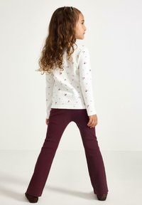 Valkoinen pitkähiha Shirt, jossa on pieniä kukkaprinttejä, yhdistetty burgundinpunaisiin samettisiin liehuviin housuihin. Lapsi, jolla on aaltoileva ruskea hius, kääntynyt sivulle.