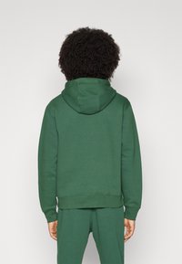 Person med lockigt hår iklädd en enkel grön hoodie och matchande byxor, som står vänd mot en ljusgrå bakgrund.