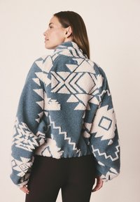 Fleecetakki sinivalkoisella geometrisella kuviolla, korkea kaulus, väljät hihat ja elastinen helma. Pehmeä texture ja mukautuva istuvuus.