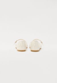 Ballet flats en cuir verni blanc et brillant avec un bout rond, une lanière arrière et un petit nœud décoratif ; semelle plate avec une finition brun clair.